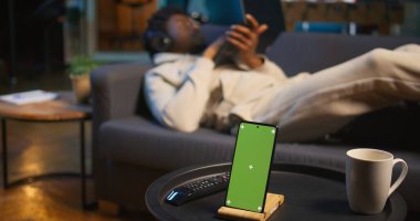 Siyah erkek uzaktan kumandalı işçi, kroma anahtar ekranının yanındaki kanepeye uzanmış, modern çatı katında, neon ışıkları ve akıllı bir televizyonla rahatlamak için boş zamanının ve boş zamanının tadını çıkarıyor. Kamera B.