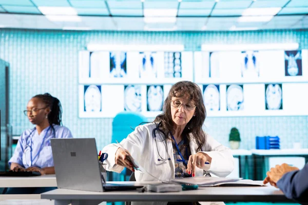 Genel pratisyen hekim röntgen taraması sonuçlarını kabinede bir erkek hastayla birlikte inceleyip muayene sırasında tarama yorumu yapıyor. Sağlık hizmetleri sağlık sigortası kapsamında.