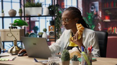 Evde klavyede yazı yazarken abur cubur yemekle meşgul. İnternetteki iş görevlerini çözmek için elektronik cihaz kullanırken kruvasan seven kadın, kamera B