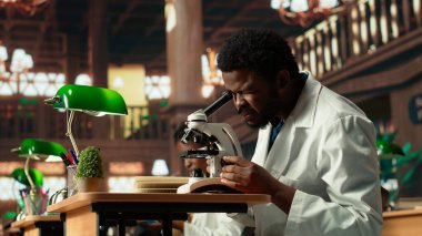 Afrikalı Amerikalı öğrenci bir kampüs kütüphanesinde mikrobiyoloji okuyor, mikroskop altında hücreleri ve mikroorganizmaları inceliyor. Erkek öğrenci biyoteknoloji tezi için hazırlanıyor. Kamera B.