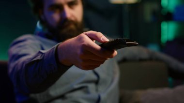 Yayınlanan içerikte kaydırmak ve film seçmek için kullanılan aygıtı kapat. Bulanık arka planda erkeğin kullandığı uzaktan kumandaya odaklan televizyon ekranında kanal değiştirme, kamera A