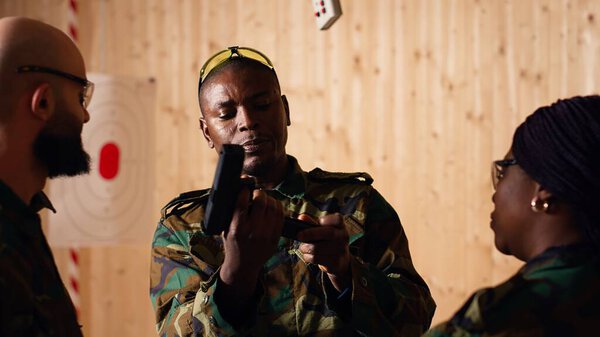 Army commander talking with recruits in shooting gallery, showing them how to reload firearms. Military officer instructing soldiers on how to load ammunition into pistol, camera B
