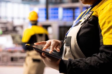 Akıllı fabrikadaki tablet ekranla etkileşime giren ellerin yakınlaştırılması, imalattaki dijital dönüşümü temsil ediyor. Yüksek teknolojili çalışma alanının arka planında bulanık güvenlik donanımlı işçi görülüyor.