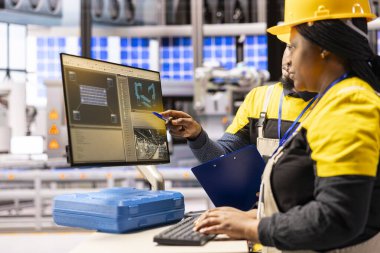 Dijital arayüzle otomatikleştirilmiş fabrika sistemini yöneten Afrikalı Amerikalı mühendisler, taşıma bantları ve montaj hatlarını optimize ediyorlar. Akıllı üretim operasyonlarını analiz eden iki profesyonel teknisyen