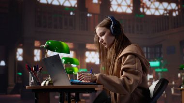 Dizüstü bilgisayarla eğitim belgeseli dinleyen ve notlar alan genç bir kız, üniversite kütüphanesinde ders çalışmak için geleneksel olmayan yöntemler kullanıyor. Bayan öğrenci veri topluyor. Kamera A.