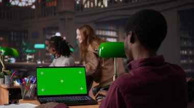 Üniversite öğrencisi, akademik kaynaklardan notlar toplarken ve bir ödev için kampüs kütüphanesi kitaplarına danışırken dizüstü bilgisayardaki bir kopyayı inceliyor. Öğrenci hayatı. Kamera B.