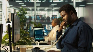 Ofisteki işçileri müşterilerle konuşmak için sabit telefonu kullanarak kapatın. İş yerindeki Afrikalı Amerikalı çalışan çağrı sırasında iş ortağıyla tartışıyor, performans ölçümlerini analiz ediyor, kamera B