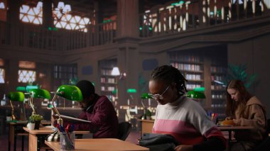 Afrikalı Amerikalı bir öğrenci kütüphane koleksiyonu veritabanındaki notlarla karşılaştırıyor ve bir edebiyat ödevine odaklanmak için dizüstü bilgisayar kullanıyor. Diploması için çalışan genç bir öğrenci. Kamera B.