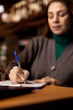 Sınıf notları üzerinde çalışan, arşivlenmiş kağıtlar ve eğitim materyalleriyle çevrili bir çalışma alanında odaklanmış bir kadın akademik bir derse hazırlanıyor, ödevlerini tamamlıyor. Kapat..