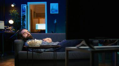 Gülümseyen adam evde kanepede uzanmış patlamış mısır yerken televizyon kanalında yayınlanan filmlere göz gezdiriyordu, işten izin almanın keyfini çıkarıyordu. Adam apartmanda uzanmış abur cubur yiyor, kamera B