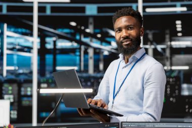 Neşeli veri merkezi mühendisinin tasviri sistem performansını dizüstü bilgisayarla izlemek, darboğazları tanımlamak. Sunucu merkezindeki iyimser adam sorun çıkarma hataları, optimizasyonlar uygulanıyor