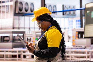 Tablet tutan odaklı fabrika müdürü endüstriyel çalışma alanındaki üretim faaliyetlerini izliyor. Siyah kadın yazılım uzmanı sistem güncellemeleri yapıyor ve depoda verimli iş akışı sağlıyor.