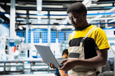 Siyah teknisyen yüksek teknoloji fabrikasında güneş enerjisi yazılım ayarları üzerinde çalışıyor. Üretim sürecini optimize ediyor. Dijital araçlar ve endüstriyel robotlarla optimal sistem yapılandırması.