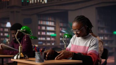 Siyahi kız kampüs kütüphanesinde sınav hazırlıklarına odaklanıyor, laptopuna huzurlu bir çalışma ortamındaki referans kitaplarından notlar alıyor. Kız genel veritabanı materyallerini kullanıyor. Kamera A.