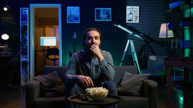 Adam evde kanepede dinleniyor, evde televizyon izlerken ağzına patlamış mısır tıkıyor. Abur cubur yiyen ve neon ışıklı oturma odasında TV şovları yapan kişi, kamera B