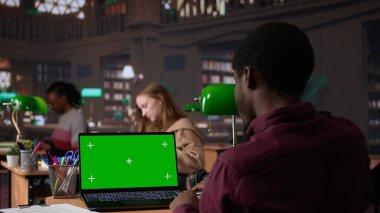 Üniversite kampüs kütüphanesinde yeşil ekranla çalışan genç öğrenci, doğrulanmış eğitim kaynaklarından akademik bilgi topluyor. Öğrenci araştırma araçlarını kullanıyor. Kamera B.