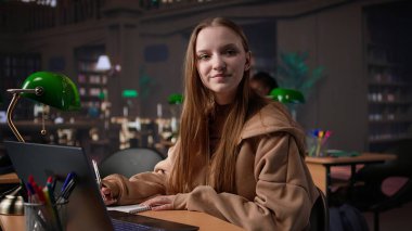 Üniversite öğrencisi sessiz bir ortamda yeşil lambanın altında dizüstü bilgisayarla çalışıyor. Önemli bir okul projesine hazırlanmak için kampüsteki üniversite kütüphanesini kullanıyor. Genç kız veri topluyor. Kamera B.