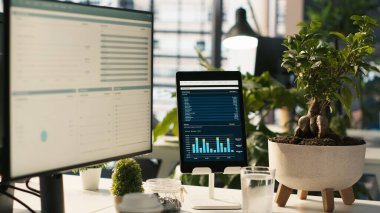 Anahtar performans göstergelerine sahip modern çalışma alanı parlak ofis ortamında saksı bitkileriyle gösteriliyor. Masanın üzerinde bilgisayar ve tablet, ekranda görünen muhasebe rakamlarıyla, görüntüyü kapat