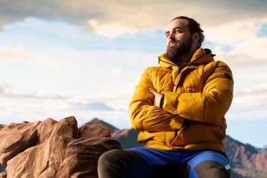 Doğa aşığı nefes kesen bir dağ sırasının kenarında duruyor, yükselen zirvelerin ve uçsuz bucaksız gökyüzünün manzaralı arka planına bakıyor. Genç adam macera ve açık hava keşiflerini seviyor..