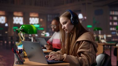 Kampüs kütüphanesinde kulaklıkla konferansa katılan genç bir öğrenci, notlar alıyor ve online bir toplantıda politika hakkında bilgi ediniyor. Ders sırasında odaklanmış bir öğrenci. Kamera A.