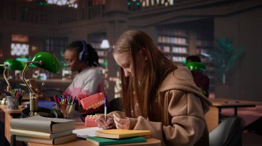 Afrika kökenli Amerikalı dişçi, insan dişlerinin plastik modelini inceliyor, kampüs kütüphanesinde stomatoloji çalışmaları yapıyor. Erkek öğrenci dişçiliği ya da oral sağlığı öğrenir. Kamera B.