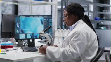 Kimya laboratuarındaki örnekleri mikroskop ve gözlem aletleriyle analiz eden siyah kadın bir uzman. Genetik araştırması, biyokimya çalışmaları ile patoloji analizi yapan bir bilim adamı. Kamera B.