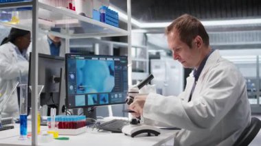 Erkek profesyonel bilim adamı, tepsi örneklerini optik mikroskop altında inceler. Büyüteçle gözlem aracı biyokimyayı ve tıbbi araştırmaları vurguluyor, biyomühendisliği. Kamera A.