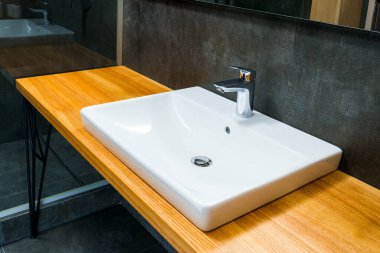 Banyoda, tezgahın ahşap yüzeyinde musluk olan beyaz seramik lavabo.
