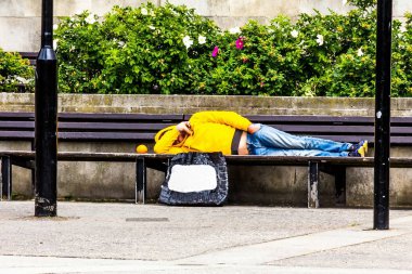 Elini yüzünü kaplayan tanınmaz işsiz kukuIeta ve kot yalan bir parkta bankta sarı ceketli