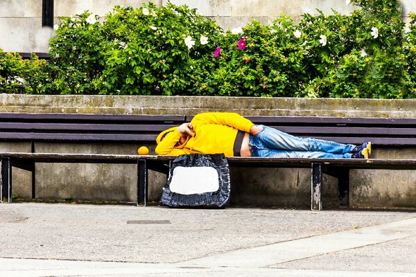 Unrecognizable unemployed in a yellow jacket with a hood and blue jeans lies on a park bench, covering his face with his hand