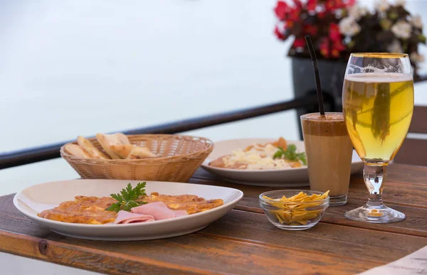 Omelet with ham and cheese served with pastry triangles, potato chips, glass of beer and coffee with straw. Morning meal set on wooden table, isolated flowers in the background.