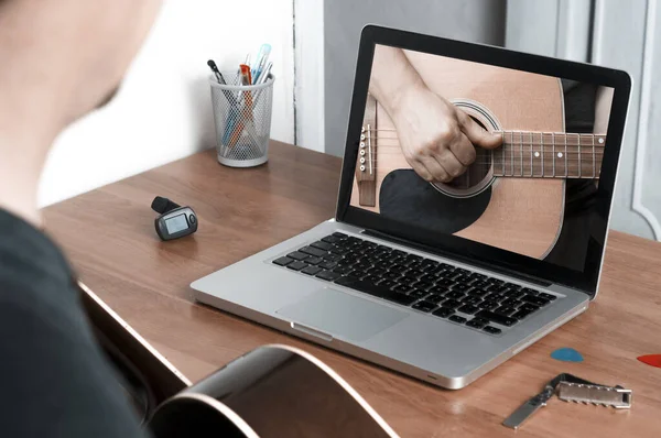 Genç adam akustik gitarlı bir dizüstü bilgisayarın önünde online gitar dersleri alıyor.