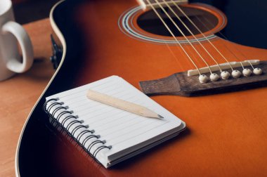 Songrwriting konsepti: arka planda bir fincan kahveyle birlikte akustik gitarın ses panosunda dinlenen bir not defteri ve bir kalem.