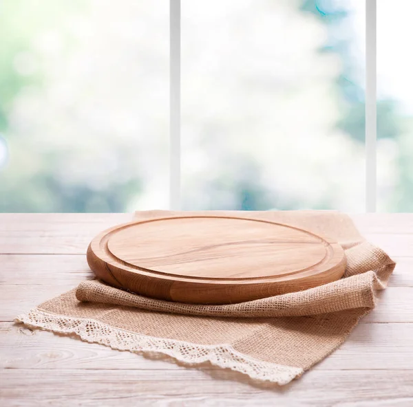 Placa de pizza de madeira texturizada vazia com guardanapo na mesa e janela da cozinha fundo borrado — Fotografia de Stock