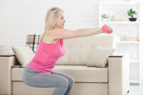 En cuclillas profundas. Vista lateral de la mujer de mediana edad en ropa deportiva haciendo sentadilla y sosteniendo pesas mientras está de pie frente a la ventana en casa . — Foto de Stock