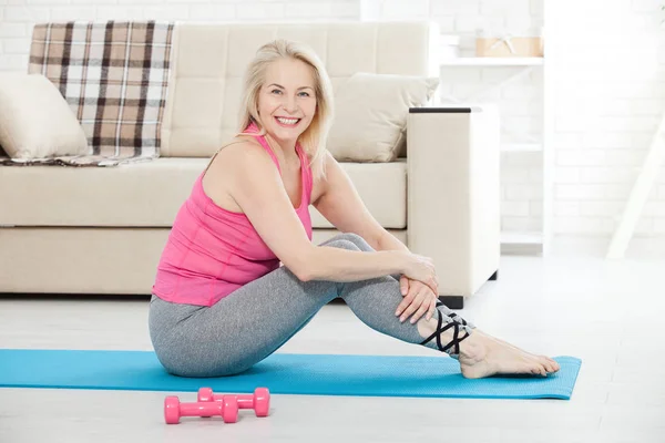 Arbeta med din kropp. Hela längden av en vacker medelålders kvinna i sportkläder, göra fitnessövningar, står framför ett fönster hemma. Realistiska bilder med sina egna brister. — Stockfoto