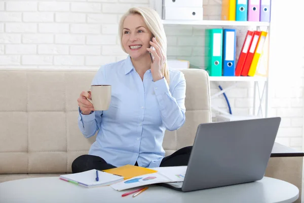 Listening to clients requirements. Beautiful middle aged woman with glasses talking on smart phone and smiling at her working place siting in the office. — Stock Photo, Image