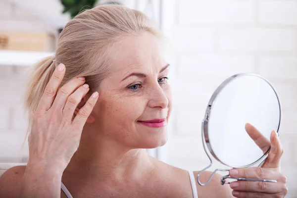 Mujer de mediana edad mirando las arrugas en el espejo. Cirugía plástica e inyecciones de colágeno. Maquillaje. Cara de macro. Enfoque selectivo — Foto de Stock