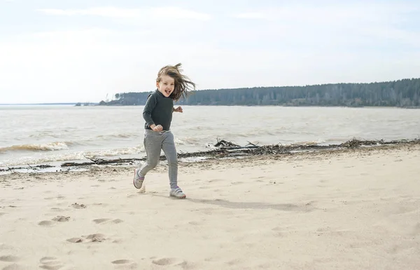 Petite fille douce aux cheveux longs bruns qui court sur la plage par une journée nuageuse. Vacances au bord de la mer. Petite fille mignonne sur la plage déserte. Summer, dehors. Du vent dans les cheveux. Paysage de plage — Photo