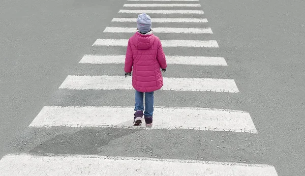 Pequeña niña linda vestida con chaqueta roja, vaqueros azules y sombrero gris cruza la calle en el cruce peatonal en el día frío. Vista superior — Foto de Stock