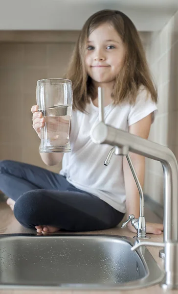 Портрет маленькой белой девочки, держащей стакан чистой воды из-под крана. Кухонный кран. Милый ребенок, наливающий пресную воду из фильтровального крана. В помещении. Концепция здоровой жизни — стоковое фото