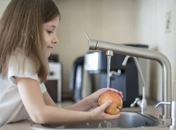 Una niña linda lava una manzana roja amarilla en un fregadero. El niño está lavando la fruta en la cocina. El niño aprende las reglas de higiene. El niño usa el grifo él mismo. Concepto de higiene de estilo de vida saludable — Foto de Stock