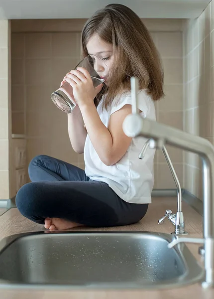 Elinde bir bardak musluk temiz su tutan küçük beyaz bir kız bebek portresi. Mutfak musluğu. Sevimli çocuk filtre musluğundan tatlı su döküyor. Kapalı. Sağlıklı yaşam kavramı