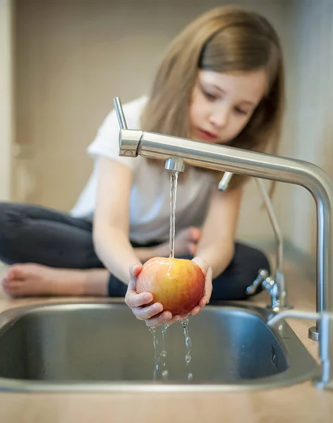 Una niña linda lava una manzana roja amarilla en un fregadero. El niño está lavando la fruta en la cocina. El niño aprende las reglas de higiene. El niño usa el grifo él mismo. Concepto de higiene de estilo de vida saludable — Foto de Stock