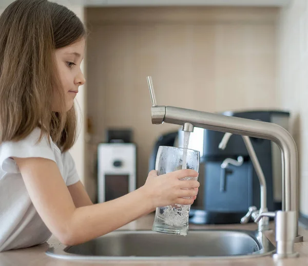 Porträtt av en liten kaukasisk flicka få ett glas kran rent vatten. Köksblandare. Söt lockig unge hälla friskt vatten från filter kranen. Inomhus. Sunt liv koncept — Stockfoto