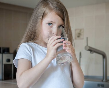 Elinde bir bardak musluk temiz su tutan küçük beyaz bir kız bebek portresi. Mutfak musluğu. Sevimli çocuk filtre musluğundan tatlı su döküyor. Kapalı. Sağlıklı yaşam kavramı