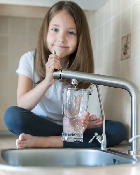 Retrato de una niña caucásica ganando un vaso de agua limpia. Grifo de la cocina. Lindo niño rizado vertiendo agua dulce del grifo del filtro. En interiores. Concepto de vida saludable — Foto de Stock
