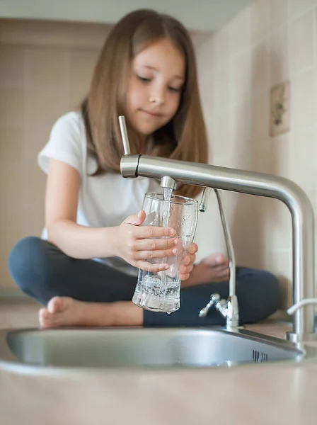 Elinde bir bardak musluk temiz su tutan küçük beyaz bir kız bebek portresi. Mutfak musluğu. Sevimli çocuk filtre musluğundan tatlı su döküyor. Kapalı. Sağlıklı yaşam kavramı