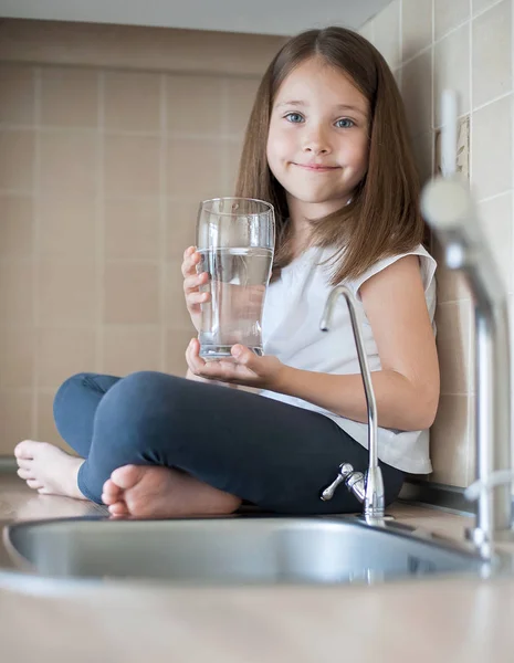 Retrato de una niña caucásica sosteniendo un vaso de agua limpia. Grifo de la cocina. Lindo niño vertiendo agua dulce del grifo del filtro. En interiores. Concepto de vida saludable — Foto de Stock
