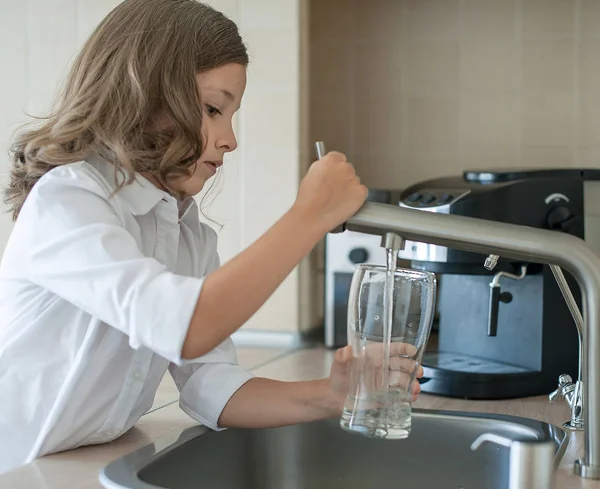 Портрет маленькой белой девочки, которая получает стакан чистой воды из-под крана. Кухонный кран. Милый кудрявый ребенок, наливающий пресную воду из фильтровального крана. В помещении. Концепция здоровой жизни — стоковое фото